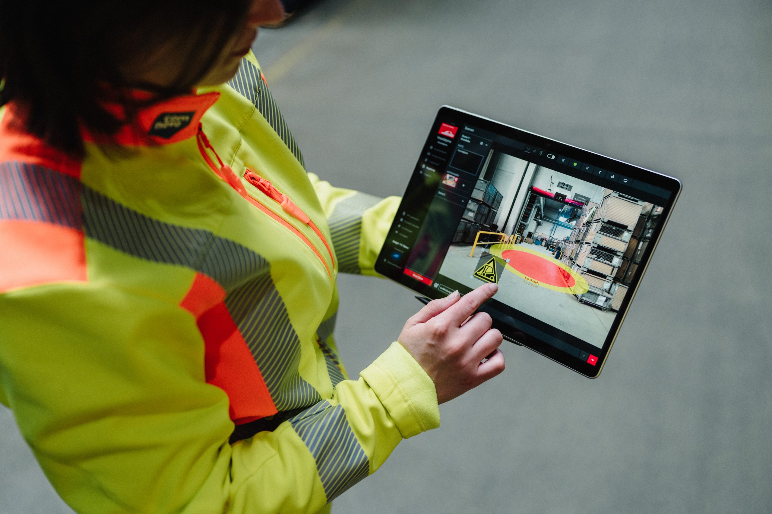 Employée en veste haute visibilité utilisant une tablette pour analyser un plan de sécurité en entrepôt avec des zones de danger.