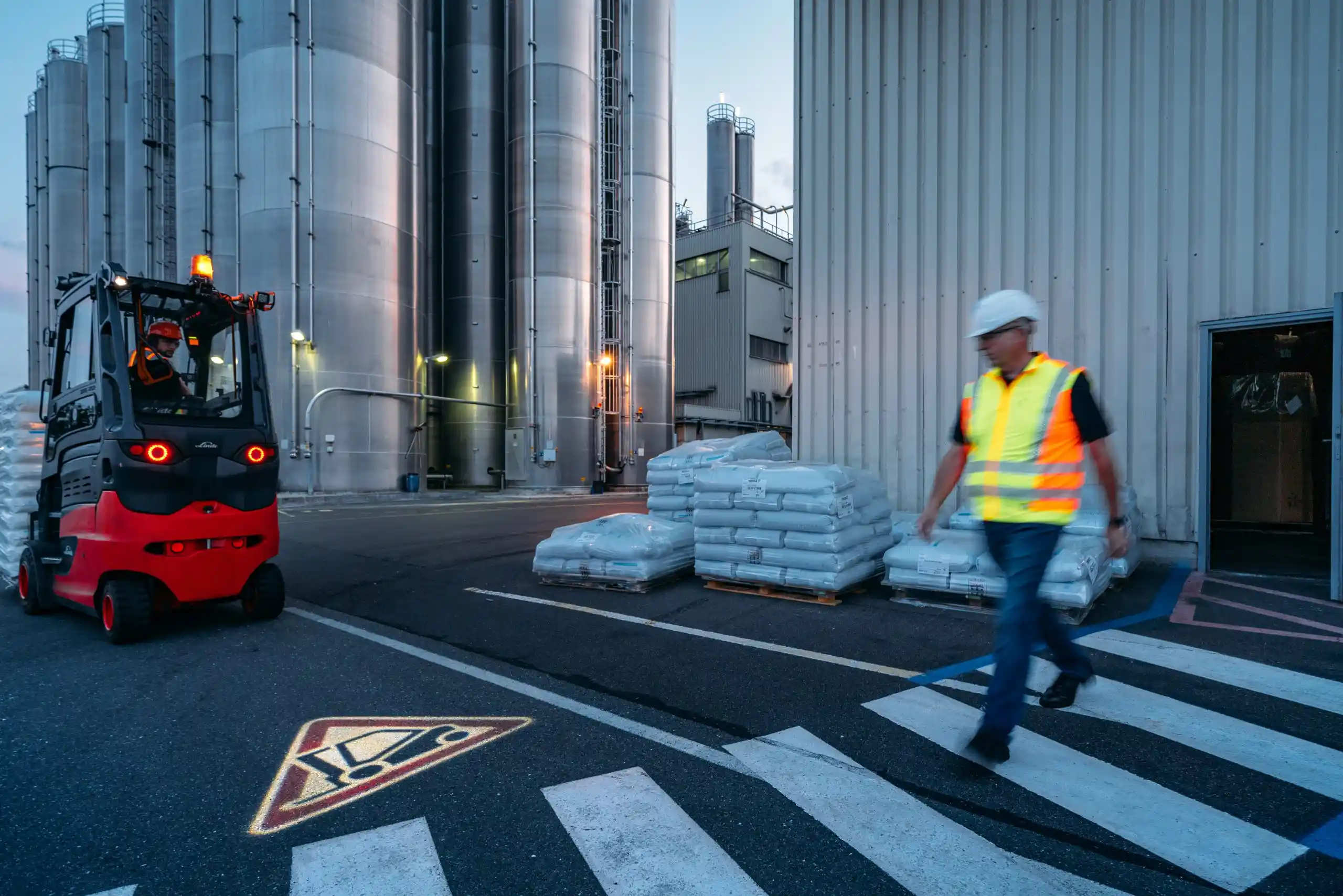 Chariot élévateur Fenwick transportant des palettes dans une zone industrielle, avec un employé en gilet de sécurité traversant un passage piéton